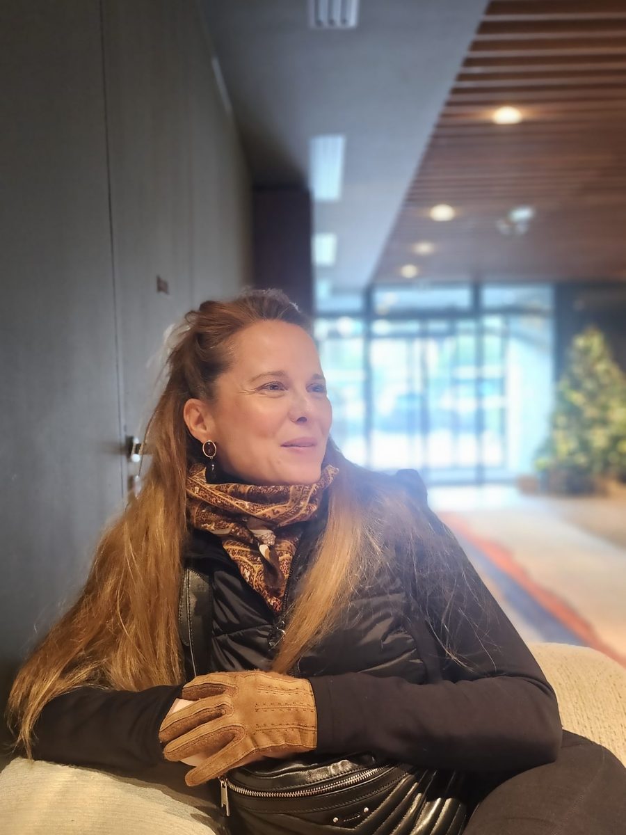 Amélie assise dans le salon de la thalasso, foulard et gants en daim, léger sourire, sapin de Noël en arrière-plan
