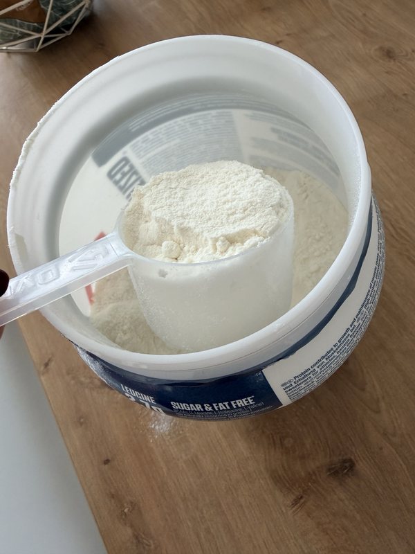 Une mesurette de whey Dymatize en poudre, tenue au-dessus du pot ouvert