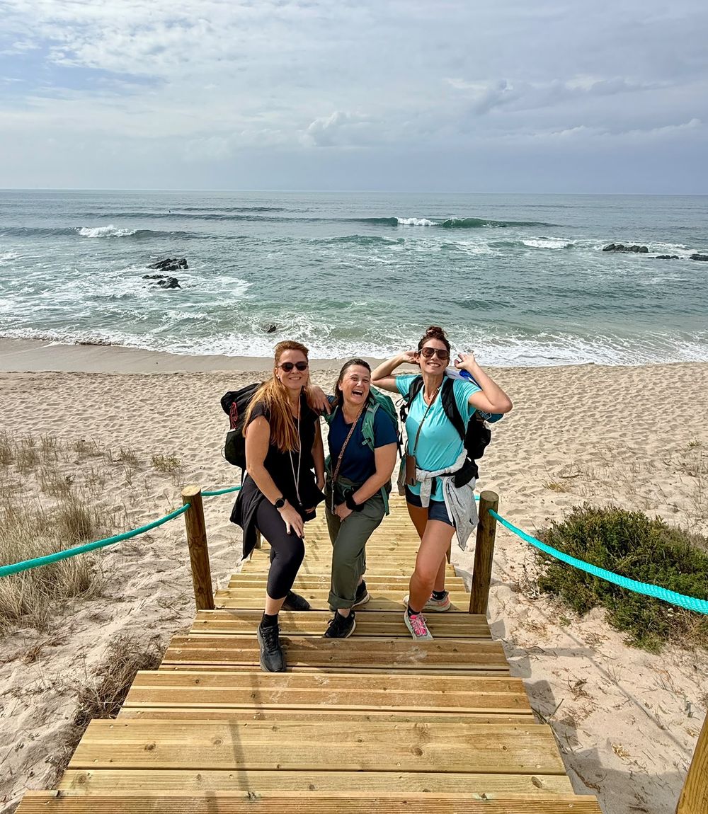 Amelie et deux amies, sac au dos, tout sourire en haut d’un escalier en bois face à la plage
