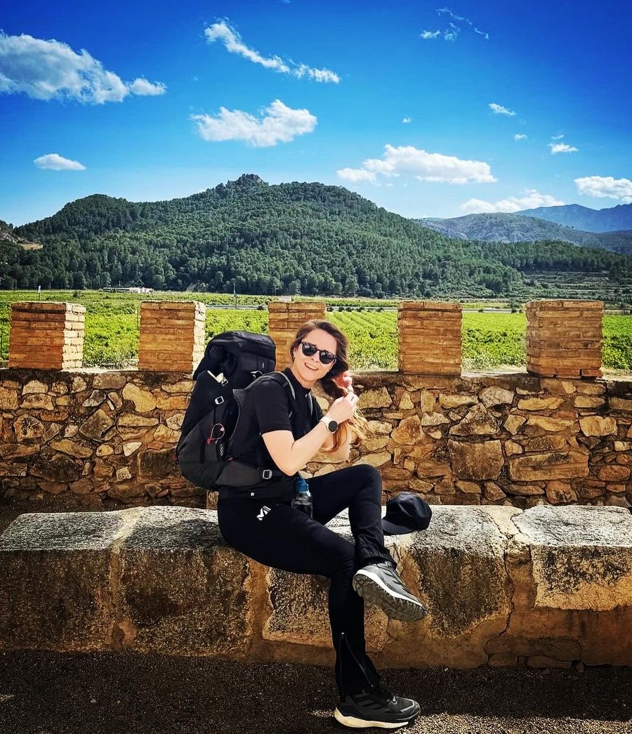 Amélie assise sur un muret de pierre, sac à dos sur l'épaule, devant les vignes et les collines