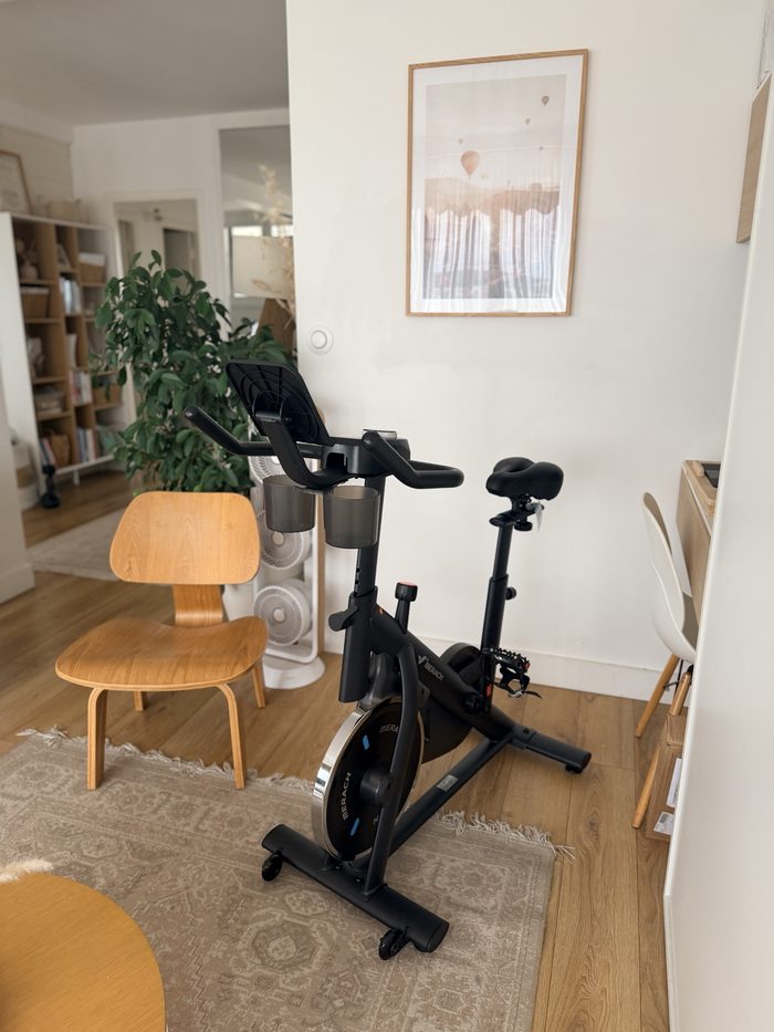 Le vélo de biking Merach dans mon salon, à côté d’une chaise en bois et d’une plante